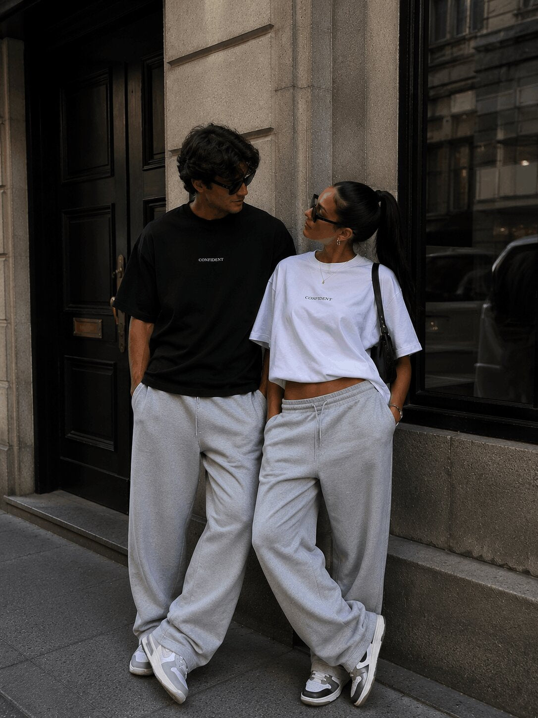 Male and female models wearing black and white CONFIDENT oversized T-shirts with grey sweatpants on a city street