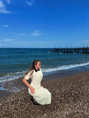 Female model seated on a pebble beach wearing a cream crochet maxi dress