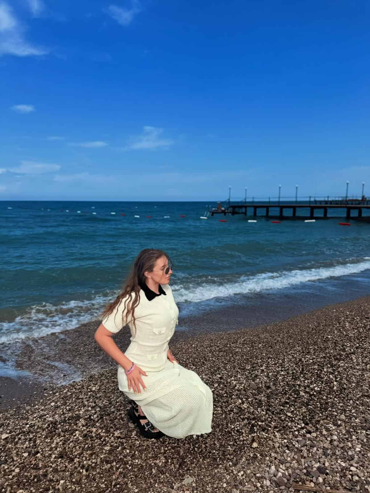Female model seated on a pebble beach wearing a cream crochet maxi dress